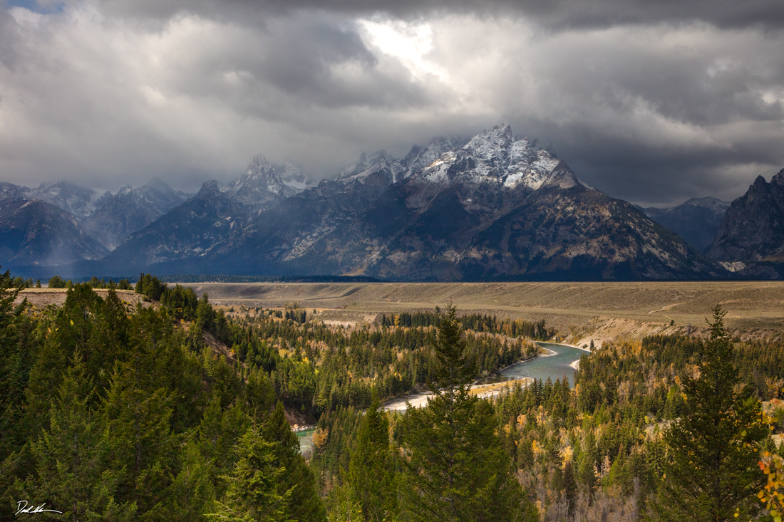 image of Grand Teton National Park during fall with pine trees in the foreground and storm clouds surrounding the mountains from Snake River Overlook