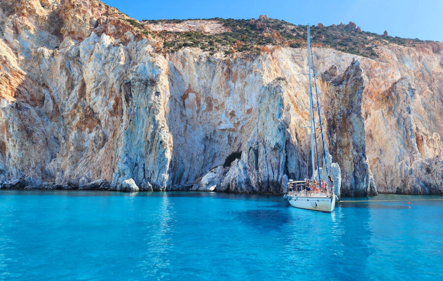 image of crystal-clear blue waters of the Aegean Sea around Greece with a sailboat in front of colorful cliffs 