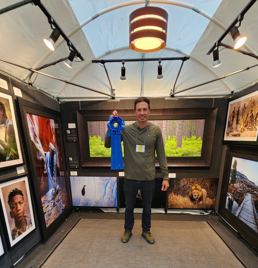 Derek Nielsen holding up his best in show ribbon at the Old Town Art Fair while standing in his booth