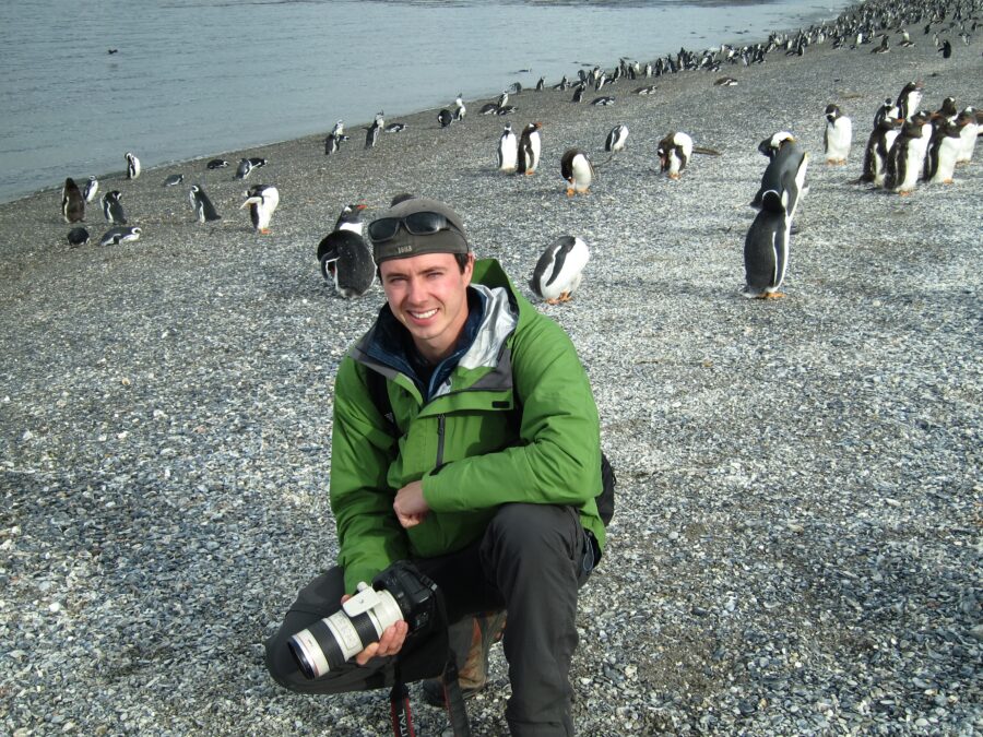 Photographer Derek Nielsen posing for a photo on a beach in patagonia surrounded by penguins early in his career